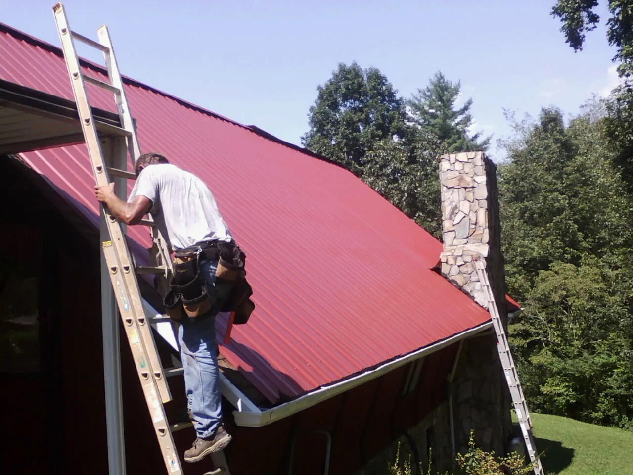 Roofer on a ladder beside a red metal roof and stone chimney — Soffit & Fascia Repair by Roofing Brothers in Kemp